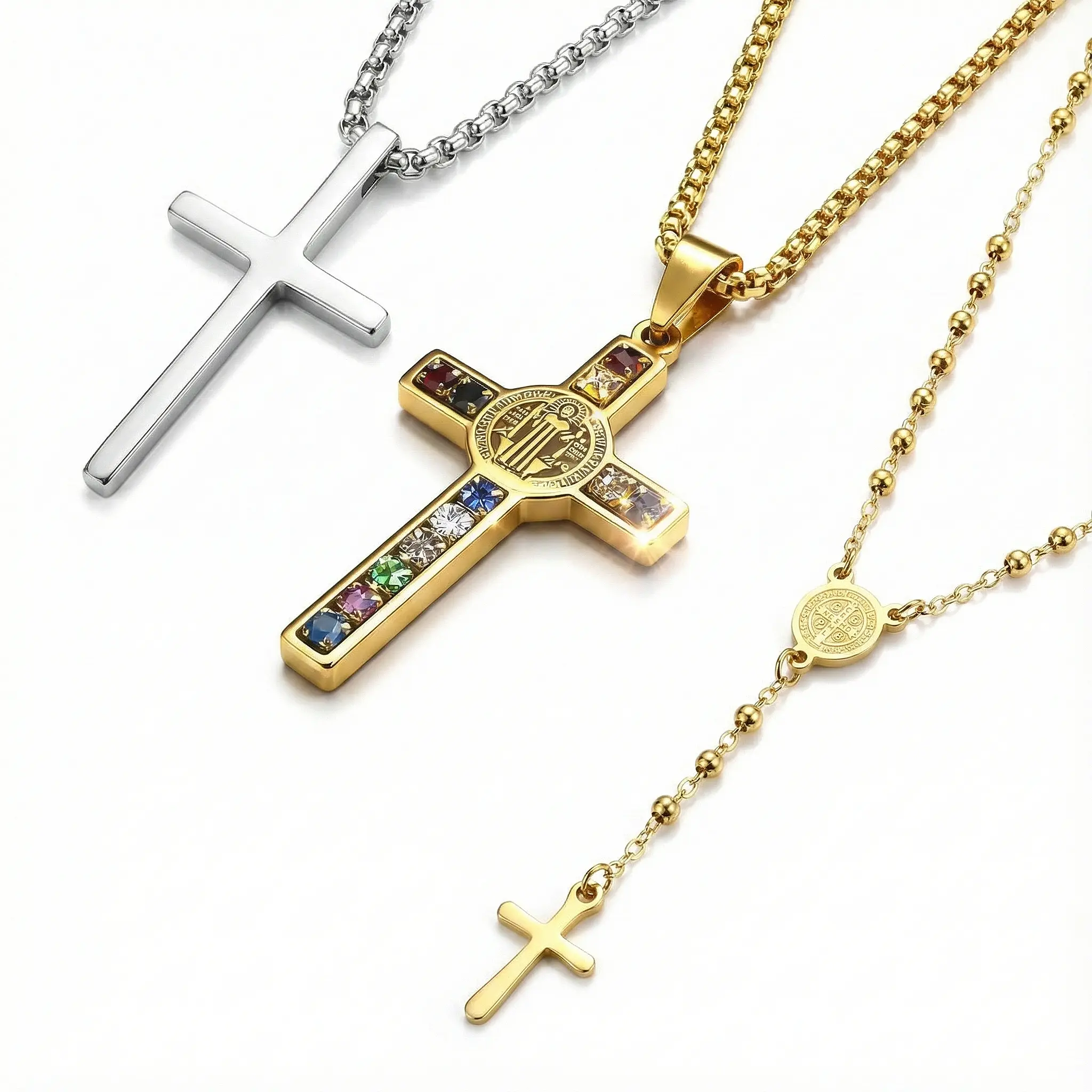 Collection of Catholic faith jewelry: A minimalist silver cross, a gold St. Benedict cross pendant with multicolor crystals, and a gold rosary bead necklace.