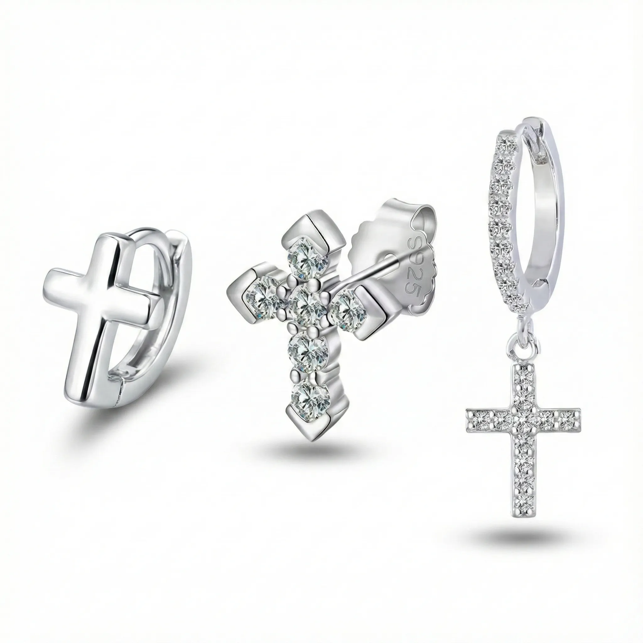 Trio of sterling silver cross earrings: Polished minimalist huggie hoop, prong-set cubic zirconia stud with visible S925 stamp, and pave crystal huggie with dangling cross charm.
