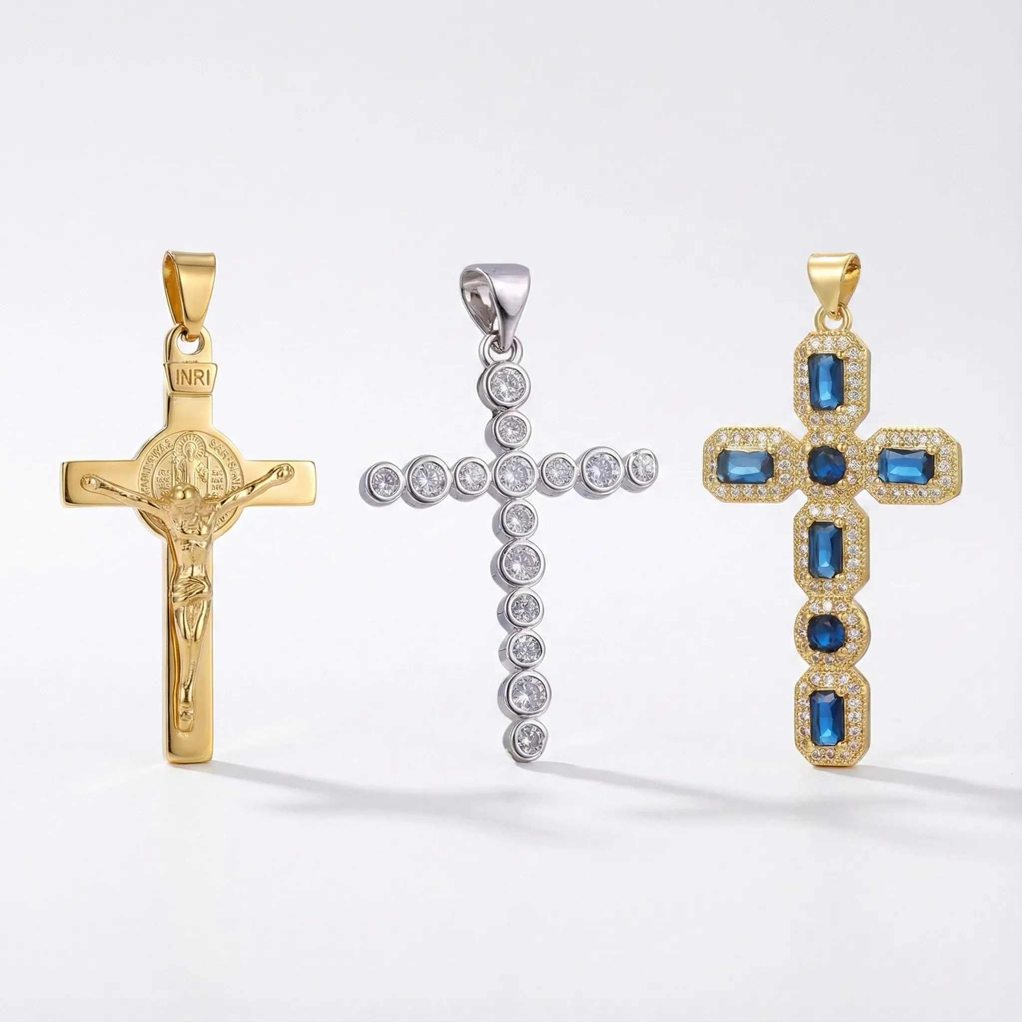 Trio of distinct cross pendants: Gold Saint Benedict Crucifix with INRI, silver bezel-set cubic zirconia cross, and vintage-style gold cross with blue sapphire crystals.