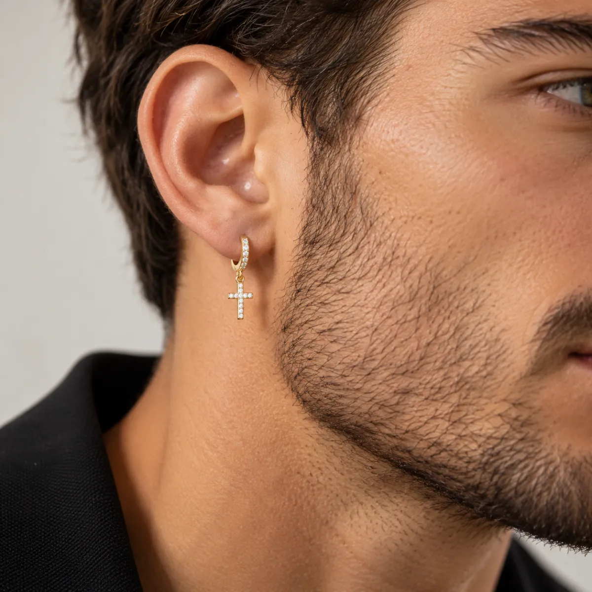 Man wearing one gold pavé cross hoop earring, close-up with beard and dark jacket