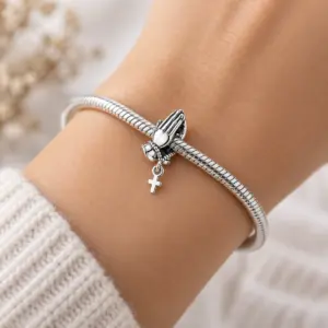 Praying hands cross charm worn on a woman's wrist with silver snake chain bracelet