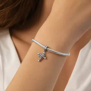 Silver cross and crown charm with pink stones worn on a womanâs wrist on a snake chain charm bracelet.