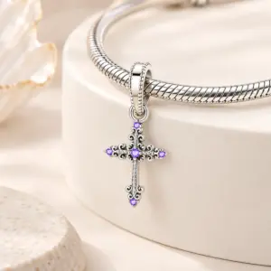 Purple stone cross dangle hanging from a silver snake chain bracelet on beige props